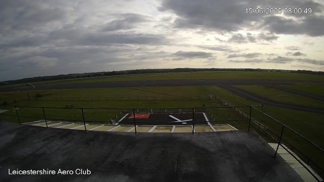 The image shows a view from a webcam at Leicestershire Aero Club, with a cloudy sky overhead. In the foreground, there is a helipad with a red and black surface, marked with a white "H" and a red square with white text. The helipad is surrounded by a fence and a railing. Beyond the helipad, there are grassy fields, taxiways, and runways at the airfield, extending into the distance under a cloudy sky. The timestamp in the top right corner indicates the date and time as 15th June 2025 at 08:00:49.