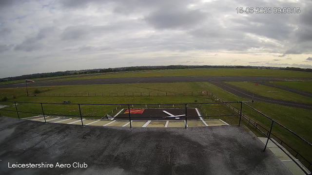 An image from a webcam showing an overcast sky above an airfield with a small windsock, grassy areas, a runway in the distance, and a parking area with a marked helipad in the foreground. The scene is labeled "Leicestershire Aero Club" in the bottom left corner, with a timestamp of 15-06-2025 09:00:15 in the top right corner.