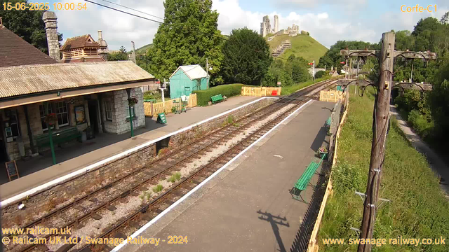 A small railway station platform with a building on the left, featuring a door, windows, and hanging flower baskets. There are green benches along the platform, and a yellow fence at the far end. A set of train tracks runs through the middle of the image, with some weeds growing between the rails. To the right, there is a wooden utility pole with wires and a shadow cast on the platform. In the background, lush green trees and grassy hills are visible, along with a hilltop castle ruin. The sky is partly cloudy with patches of blue. The date and time displayed are June 15, 2025, at 10:00:54.