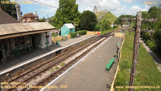 A small railway station platform with a stone building to the left, featuring a tiled roof and a chimney. The platform has a green bench, a couple of people sitting and chatting near the benches, and a small green shed behind them. In the distance, there are lush green trees and a hill with a castle ruin on top. Overhead, electrical wires run parallel to the train tracks, which extend into the distance. The sky is partly cloudy, with patches of blue visible. There are some flower pots on the fence along the platform.