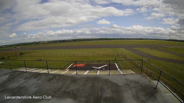A view from a webcam at Leicesterhire Aero Club showing an outdoor aircraft parking area with a large white and red "X" marking on the ground. In the background, there is a runway with green fields beyond, under a partly cloudy sky. The time and date stamp in the top right corner reads 15-06-2025 11:59:52. The bottom left corner has the text "Leicestershire Aero Club."