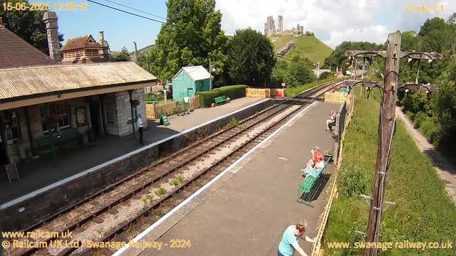 A small train station with a platform, several passengers sitting on benches, and a railway track running through the scene. In the background, there is a hill with ruins of an old castle on top. On the left side, there is a building with a tiled roof and a person standing near the entrance. A green shed and some greenery are also visible near the platform. The sky is partly cloudy with some sunlight. There are electrical poles and wires above the tracks. The scene appears calm and quiet.