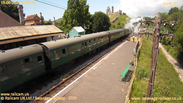 A vintage green train is stopped at a small railway station, emitting white steam from its engine. The station platform is empty except for three people standing near the train. To the right, there are old wooden electrical poles with multiple wires and insulators. In the background, there is a lush green hillside with ruins of an ancient castle on top, and a clear blue sky with some clouds. To the left, there is a building with a tiled roof and chimney. The time and date displayed on the image are June 15, 2025, at 13:00:42.