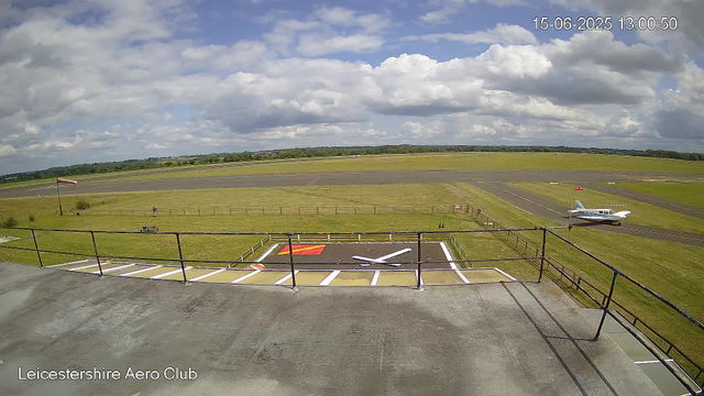 An outdoor view at Leicestershire Aero Club showing a runway under a partly cloudy sky, with a small airplane parked on the grass near the runway. In the foreground, there is a helipad with a white "H" marked on it, surrounded by a metal railing. The grass areas are green, and the horizon features a line of trees. The timestamp indicates the date and time as 15th June 2025 at 1:00 PM.