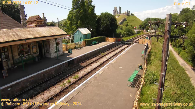 A small railway station platform with a stone building to the left, featuring a tiled roof, window boxes, and a bench underneath a lamppost. There is a small green shed and a green bench behind it. The tracks run parallel to the platform, with a path and greenery on the right side. In the background, a grassy hill with castles ruins sits atop, under a partly cloudy sky. Several utility poles with wires run alongside the tracks. The date and time are displayed at the top left, and website information appears at the bottom.