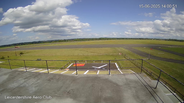 A view from a webcam showing a runway and adjacent grassy areas under a partly cloudy sky. There is a windsock on the left side, some parking markings in the foreground, and a small section of fenced-off grass near the runway. The image is timestamped "15-06-2025 14:00:51" and has the label "Leicestershire Aero Club" at the bottom.