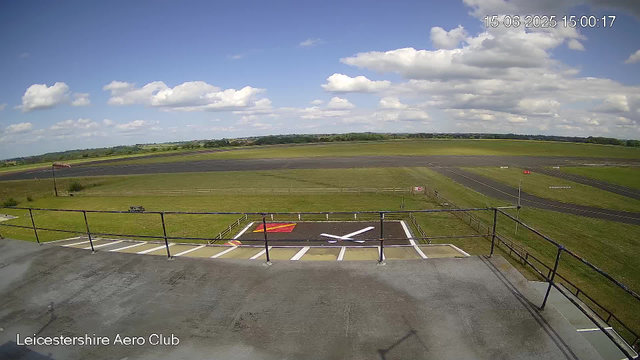 An outdoor view from a webcam at Leicesterhire Aero Club showing a clear sky with scattered clouds, an airstrip in the distance, and a helipad with a white and red marking in the foreground. The scene is bright and appears to be daytime. There is a railing and a platform area at the bottom of the image. The timestamp on the image reads 15-06-2025 15:00:17.