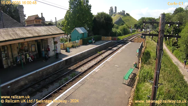 A small train station platform with a stone station building on the left, where a few people are sitting or standing. There are some benches, a blue sign, and a small green shed behind the platform. On the right, there are old wooden railway poles with electrical wires, alongside a narrow path bordered by greenery. In the background, there is a hill with an ancient castle ruin on top, and a lush landscape with trees and a clear sky. The timestamp indicates the date and time as June 15, 2025, at 16:00:48.
