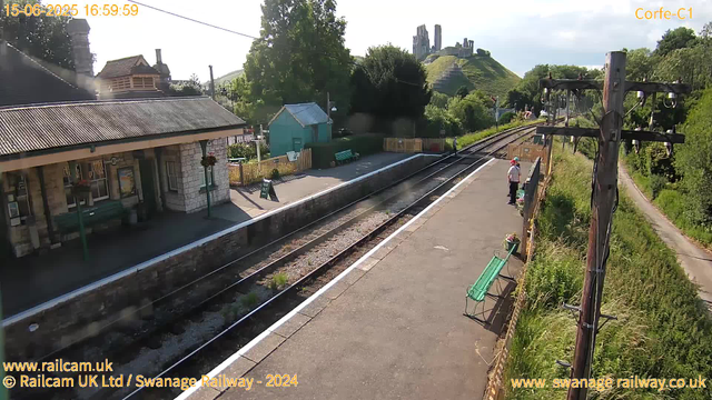 A small train station platform with a stone building on the left, featuring green benches and hanging flower baskets. There are train tracks running through the center, with a person wearing a white shirt, dark pants, and a red cap standing on the right side of the platform. In the background, green trees, a small teal-colored building, a hill with ruins on top, and a clear sky are visible. Overhead, power lines run across the scene. The timestamp on the image reads 15-06-2025 at 16:59:59.