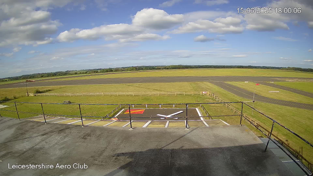 A view from a webcam shows a runway and open field at Leicestershire Aero Club under a blue sky with scattered clouds. In the foreground, there is a concrete platform with black metal railings and painted markings on the ground. There's an empty helipad designated with a large white "H" in the middle, a red square, and a white arrow pointing to the right. The landscape extends into a grassy area with a fence, and beyond that, the runway with dark asphalt and white markings. In the distance, there are trees and fields. The timestamp in the top right corner reads "15-06-2025 18:00:06."