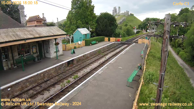 A small train station platform with a stone building on the left, green benches, potted plants, and a yellow fence lining the edge of the platform. Railroad tracks run parallel to the platform, with overhead electrical wires and poles. In the background, a green hill with a castle ruins perched on top, surrounded by trees and a cloudy sky. There is a footpath on the right running alongside the tracks, bordered by grass and shrubs. The timestamp indicates the date and time as June 15, 2025, at 6:00 PM.