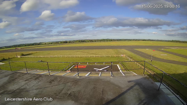 A wide view of an airfield from a webcam, showing a small, fenced tarmac area with a landing strip in the background under a partly cloudy sky. The clock overlay shows the date as June 15, 2025, and the time as 19:00:16. The location label reads "Leicestershire Aero Club."
