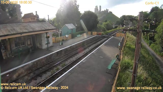 A small railway station platform with a stone building on the left side, and a train track running through the center. There are two green benches along the platform, and a wooden fence borders the area. In the background, there are trees, a small teal-colored shed, and a large hill with a historic castle or ruins on top. An old railway telegraph pole is on the right side, with wires hanging from it. The sky is partly cloudy with the sun low, creating bright sunlight and some lens flare. The timestamp shows the date as 15th June 2025 at 19:00:30.