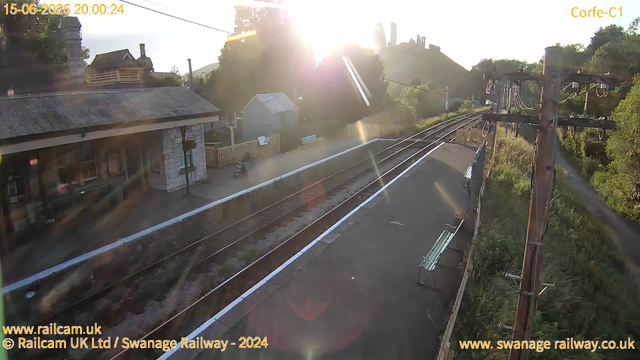 A small train station platform is shown in the image, with the sun setting behind hills in the distance. The platform has a bench and a small shelter. Adjacent to the platform are railway tracks with a white safety line along the edge. There is an electrical pole with wires on the right side, and trees and greenery surrounding the area. The scene appears calm and quiet, with no trains present. The timestamp indicates the time as 20:00:24 on June 15, 2025, and the location is Swanage Railway.
