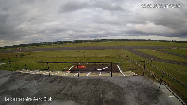 The image shows a view from a webcam at Leicesterhire Aero Club, overlooking an empty helipad with a large X marking the landing area. The surroundings include grassy fields, a paved runway, and a cloudy sky overhead. The scene appears to be captured in the evening, as indicated by the timestamp.