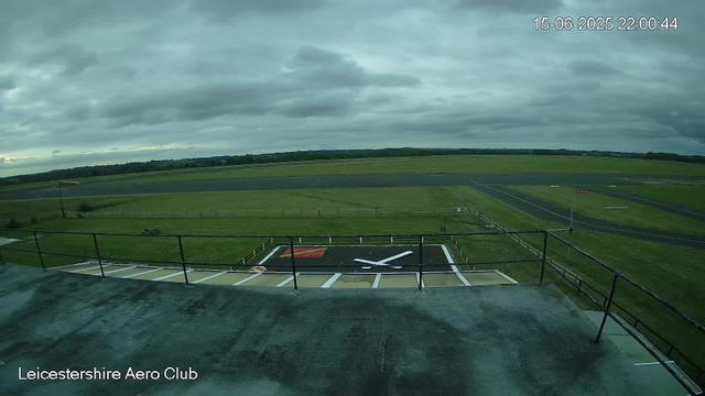An image from a webcam at Leicesterhire Aero Club shows a cloudy sky over a grassy airfield. In the foreground, there is a helipad with a large white helicopter tail rotor symbol painted on it, and a red safety box nearby. The airstrip extends into the distance, bordered by green fields and a paved runway. The timestamp shows the date and time as 15-06-2025 at 22:00:44.