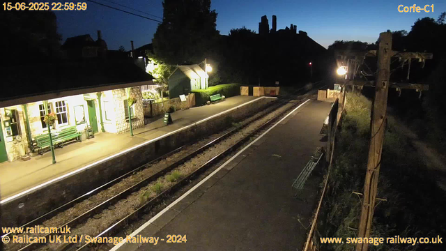 A railway platform at dusk, with a small stone building illuminated by outdoor lights on the left. The platform has green benches and flower baskets hanging beneath the windows. There is a waste bin and a small signboard near the building. The railway tracks run down the center, and the far end of the platform is bordered by a wooden fence with a green gate. In the background, dark silhouettes of trees and a hill with some structures or ruins are visible against the early night sky. A wooden utility pole with wires is on the right, with a dimly lit path running alongside it. The timestamp on the image shows the date and time as 15-06-2025 at 22:59:59.