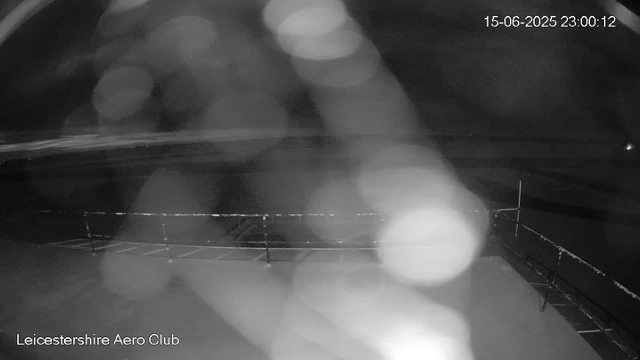 A nighttime view from a webcam at Leicestershire Aero Club shows a dark sky with few visible details. There are blurry lights and reflections, with the timestamp indicating it was taken on June 15th, 2025, at 23:00:12. The scene appears to include part of a rooftop or deck area with a railing visible at the bottom of the image. The overall image is hazy and low-contrast.
