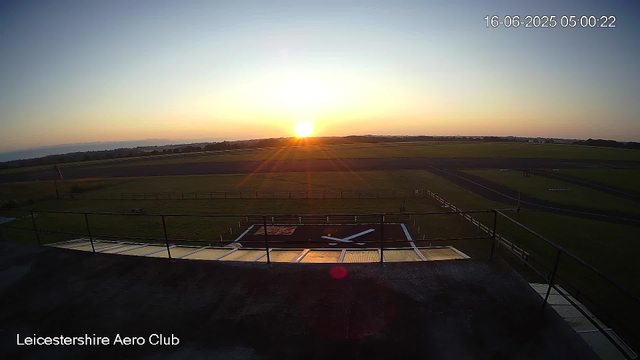 The image shows a sunrise over an open grassy field at Leicestershire Aero Club. The sky is mostly clear with a few clouds on the horizon, and the sun is just above the horizon, casting a warm glow across the landscape. There is a helipad in the foreground with a large white "H" painted on it, surrounded by a safety railing. The date and time are displayed in the top right corner as 16-06-2025 05:00:22.