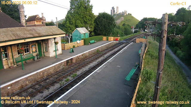 A small, quaint railway station platform with a stone building featuring green benches and a green door. There are hanging flower baskets on the building. The platform has a few green benches, a sign saying "WAY OUT," and some small potted plants. Two benches are visible on the right side of the platform near a wooden fence. Overhead, there are tall wooden poles supporting electrical wires for the train. In the background, lush green trees and a hillside with an ancient castle ruin are visible, along with a green garden shed next to the platform. The scene is bright, indicating early morning sunlight. The date and time at the top left indicate it was captured on June 16, 2025, at 6:00 am.