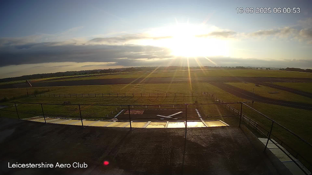 A sunrise over an open grassy field with a runway or track, seen from a webcam at Leicestershire Aero Club. The sky has some clouds, and the sunlight creates a bright glare. The timestamp in the top right corner shows the date and time as 16-06-2025 at 06:00:53.