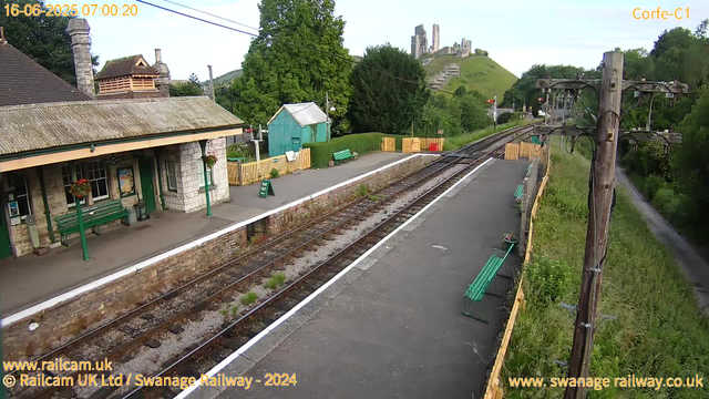 A small, rural train station platform with a building on the left side, decorated with hanging flower baskets. There are green benches on the platform, and a yellow fence borders the area. The railway tracks run parallel to the platform, curving slightly in the distance. Overhead electric lines are supported by wooden poles. In the background, lush green trees and a grassy hill topped by ancient castle ruins are visible. The sky is clear with a few clouds, and the scene is brightly lit, indicating early morning.