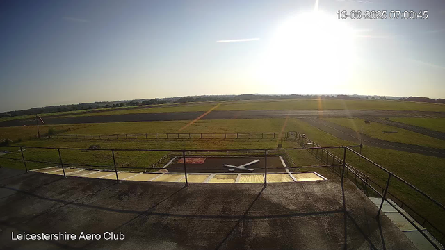 A view from a webcam at Leicesterhire Aero Club showing a bright, sunny sky with minimal clouds. The scene includes a runway stretching into the distance, grassy fields on either side, and a windsock on the left. The time and date are displayed in the top right corner as 16-06-2025 07:00:45. The foreground shows part of a rooftop with a weather vane or anemometer on it. The bottom of the image has the text "Leicestershire Aero Club."