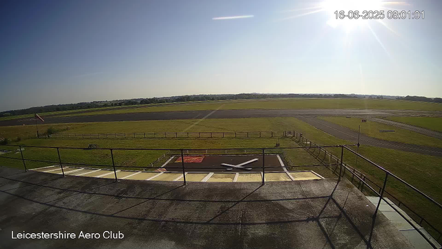 The image shows an outdoor scene at Leicestershire Aero Club during daytime. In the foreground, there's an aircraft parked on a tarmac area, viewed from above. The aircraft is facing left and appears to be a small, light aircraft. Surrounding the tarmac, there is a grassy area with a wooden fence. Beyond that, the scene extends into open fields and a runway, with clear skies, bright sunlight, and the date and time displayed as June 16, 2025, at 8:01 AM. The sun is shining brightly from the upper right corner of the image.