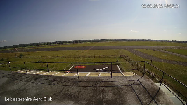 A view from a webcam at Leicestershire Aero Club showing the open tarmac and grassy areas of an airfield under a clear sky. The foreground features a large clock on a platform with the time as approximately 9:09, and the image is timestamped 16-06-2025 09:00:21. The horizon shows distant trees and open fields.
