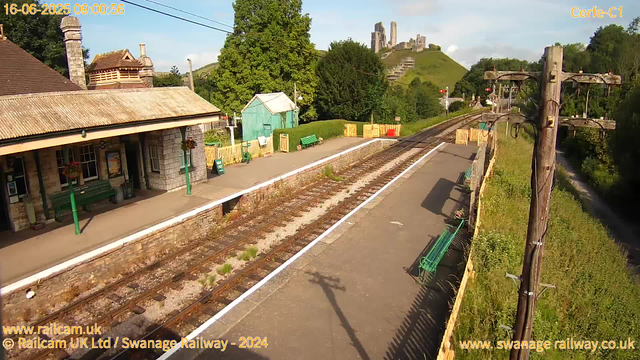 A small railway station platform with a building on the left, decorated with hanging flower baskets. Several green benches are placed along the platform. The railway tracks run parallel to the platform, with some plants growing between the sleepers. On the right side, there is a wooden fence with electrification poles and wires running above the tracks. In the background, lush green trees cover rolling hills, with an ancient castle ruin visible on top of a hill in the distance. The sky is mostly clear with a few clouds, and the image is timestamped June 16, 2025, at 9:00 AM.