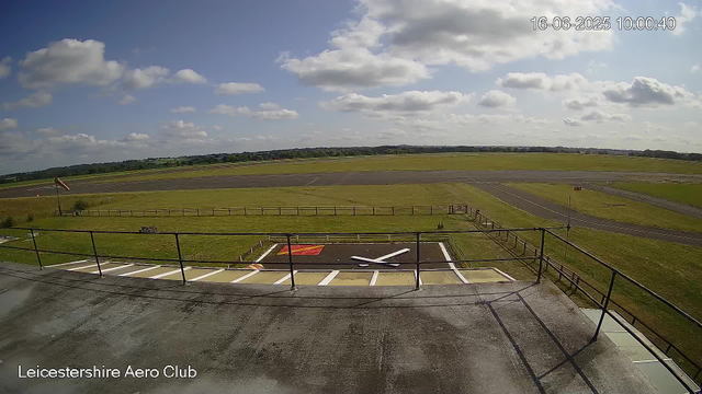 A wide view of an airfield taken from a webcam, showing a runway in the distance under a partly cloudy sky. In the foreground, there is a rooftop with a helipad marking white lines and a red rectangle, a small windsock on a pole, and a wooden railing. The image is labeled "Leicestershire Aero Club" at the bottom left corner, and the date and time are displayed in the top right corner as "16-06-2025 10:00:40".