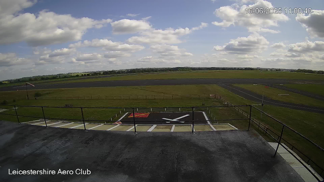A photo taken from a webcam at Leicester Aero Club shows the sky with scattered clouds over an open airstrip. In the foreground, there is a helicopter landing pad with a large white "X" on it, bordered by black and white lines. The surrounding area is grassy with a fence and a small windsock to the left. In the background, there are long, flat runways and open fields stretching towards the horizon. The timestamp on the image reads 16-06-2025 11:00:40.