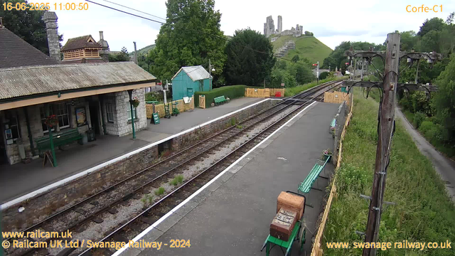 A small train station platform with a stone building on the left, featuring a sloped roof, windows, and benches underneath the shelter. There are green benches and a small green shed on the platform. A yellow wooden fence surrounds part of the station, and there are potted flowers and plants along the edge. Railway tracks run through the middle, with greenery and trees on both sides. In the background, a lush green hill with the remains of a historic castle or ruins atop it can be seen under a partly cloudy sky. An overhead electrical line runs parallel to the tracks, supported by tall wooden poles.