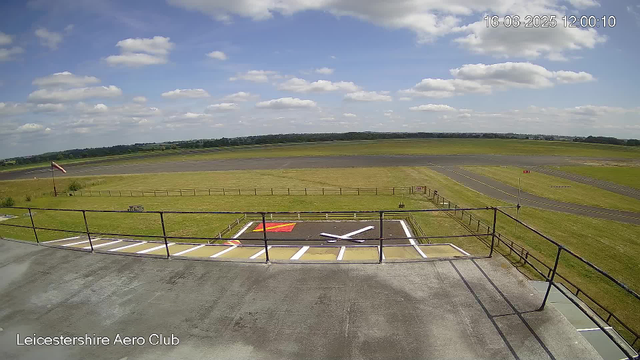 The image shows a clear view from a webcam at Leicestershire Aero Club on a bright, sunny day with a partly cloudy sky. In the foreground, there is a small paved area with parking spaces marked and a designated helipad painted with a large white "H" and a nearby red square. Metal railing surrounds the parking area, and a flagpole with a flag waving in the breeze can be seen on the left side. Beyond this, grassy fields and an aircraft runway stretch into the distance, with some runway markings visible. The timestamp in the top right corner indicates the photo was taken on 16th June 2025 at 12:00:10. The location information is displayed at the bottom left as "Leicestershire Aero Club."