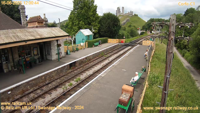 A small train station platform with a stone station building on the left and a row of green benches on the right. A few older people are sitting on the benches, and a woman with a dog is walking near a small turquoise shed in the background. There are railway tracks running through the scene, with a grassy hill and castle ruins on a distant hill in the background. An old wooden pole with electrical wires runs along the right side of the platform. The timestamp shows the date and time as 16-06-2025 at 12:00:44. The sky is partly cloudy with patches of sunlight.