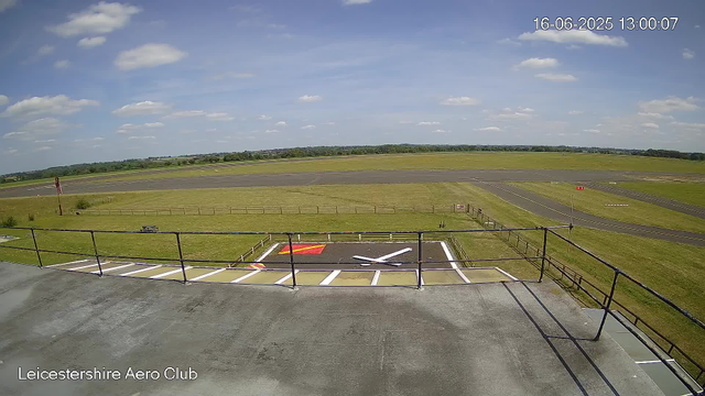 A clear view from a webcam at Leicestershire Aero Club shows a runway and open sky. In the foreground, there is a parking area with painted markings and a wind sock. The sky is partly cloudy with fluffy white clouds scattered across a blue sky. The landscape is flat and stretches into the distance with green fields and trees on the horizon. The timestamp indicates the date and time as June 16, 2025, at 1:00 PM.