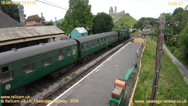 A vintage green passenger train is stopped at a small railway station. The station platform has green benches, potted plants, and a wooden luggage cart. To the right, there are wooden electrical poles with wires. In the background, there is a lush green hillside with a weathered castle ruin on top. To the left, there are traditional buildings with tiled roofs and a small turquoise shed. Beyond the train, dense trees and a pathway run parallel to the tracks. The timestamp at the top left reads "16-06-2025 13:01:02," and the top right displays "Corfe-C1."