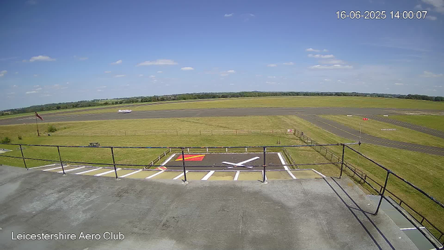A runway at Leicestershire Aero Club under a clear blue sky with scattered clouds. An airplane is taxiing on the runway in the distance. In the foreground, there is a small parking area with marked spaces, some of which are empty, and a black railing along the edge of a balcony or viewing platform. A windsock is visible on the left side of the image. The timestamp in the top right corner indicates the date and time as 16th June 2025, 14:00:07.