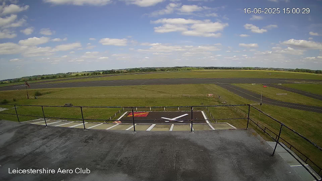 A view from a webcam at Leicestershire Aero Club showing a clear blue sky with scattered white clouds. In the foreground, there is a helicopter landing pad with a large white "H" and a red marking. The surrounding area is grassy, with a fence around the landing pad and a paved observation deck or platform at the top of the image. In the distance, there are open fields and a few pathways or runways. The timestamp in the top right corner reads "16-06-2025 15:00:29."