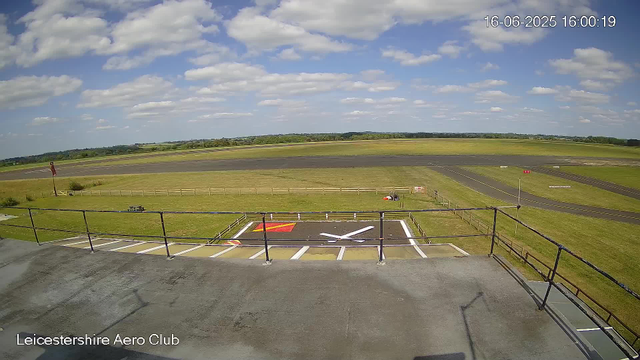 A wide view of an airfield with a runway in the distance under a partly cloudy sky. In the foreground, there is a helipad with a large white "H" and red markings, surrounded by a small fence. There is a grassy area beyond the helipad with a few small objects and a flagpole on the left side. The timestamp in the top right corner reads "16-06-2025 16:00:19," and the text at the bottom left reads "Leicestershire Aero Club."