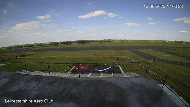 An outdoor view from a webcam at Leicestershire Aero Club shows a small helicopter landing pad with a large white helicopter silhouette painted on it. The sky is partly cloudy with a few scattered clouds, and the landscape includes green fields, a runway in the distance, and a flagpole with a flag on the left side. The timestamp in the top right corner indicates the date as June 16, 2025, and the time as 17:00:28. The bottom left corner displays the location as "Leicestershire Aero Club".