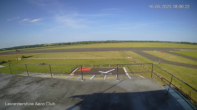A view from a webcam at Leicesterhire Aero Club shows a clear blue sky with a few small clouds, a runway in the distance, and a small area with painted markings and a white directional arrow on the ground in the foreground. There is a grassy area surrounded by a white fence, a waving windsock on a pole, and some parking or taxiway markings on the pavement. The image is timestamped June 16, 2025, at 6:00 PM.