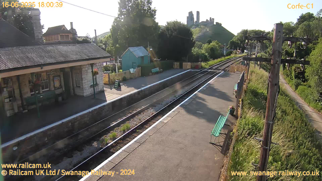 A small train station with a platform on the left side and railway tracks running through the center. There is a building with a tiled roof, benches, and a small blue shed near the station. To the right, there are weathered wooden utility poles and green benches along the edge of the platform. In the background, there are trees, greenery, and a hill with what appears to be castle ruins or old ruins on top. The sky is clear and sunny. The timestamp in the top left corner indicates the date and time as June 16, 2025, at 6:00:39 PM.