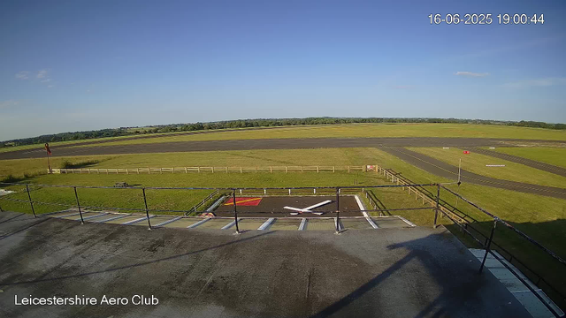 A view from a webcam overlooking an airfield with a clear blue sky. In the foreground, there is a raised platform with a safety railing and a large clock face lying flat on the ground. The airfield has grassy areas, runways, and some small obstacles. The date and time display in the top right corner reads "16-06-2025 19:00:44." The text "Leicestershire Aero Club" appears in the bottom left corner.