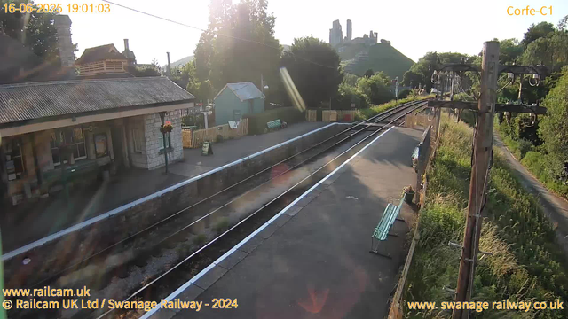 A small train station platform is visible with a building on the left side, adorned with hanging flower baskets. Multiple railway tracks run through the scene, with the sun low in the sky, casting bright light and long shadows. There are two benches along the platform, and a small structure or shed in the distance. To the right, power lines and electrical poles run parallel to the tracks. In the background, there is a green hillside with a castle or fort ruins atop it, partially illuminated by the sunlight. The scene appears to be early evening, with the timestamp indicating 19:01 on June 16, 2025.
