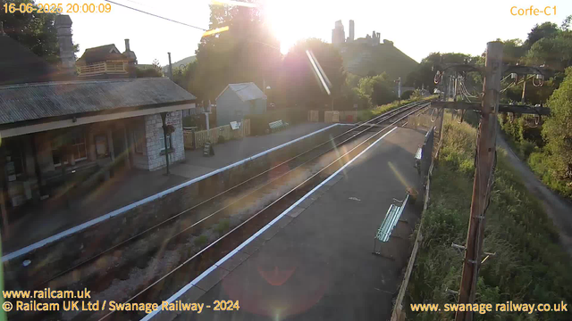 A railway platform with a building on the left, with the sun setting behind trees and hills in the background. There are empty benches on the platform, and a railway track runs along the right side. Electric lines and poles run parallel to the tracks, and some greenery is visible along the right side. The scene appears to be late evening.