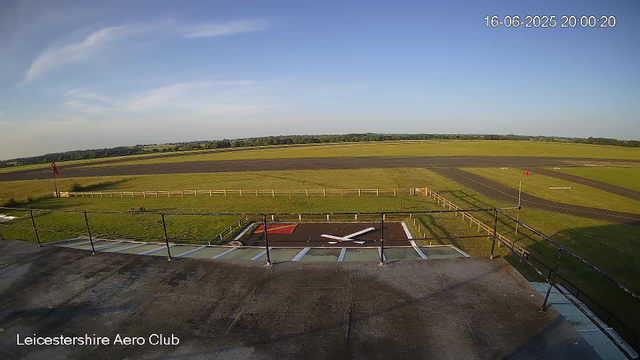 An empty aircraft parking area with a single helicopter landing pad at Leicesterhire Aero Club. The control tower's balcony is in the foreground, with a small helicopter on a pad below. Beyond are open grassy fields and a clear blue sky. The timestamp shows the date and time as June 16, 2025, at 8:00 PM.