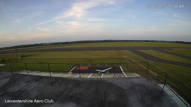 A wide view of an airfield at sunset, showing runways, grassy areas, and a clock on the ground resembling an airplane propeller. The sky is partly cloudy with soft light, and the timestamp in the upper right corner reads "16-06-2025 21:00:49." The enclosure is marked with a railing, and the location "Leicestershire Aero Club" is displayed in the bottom left corner.