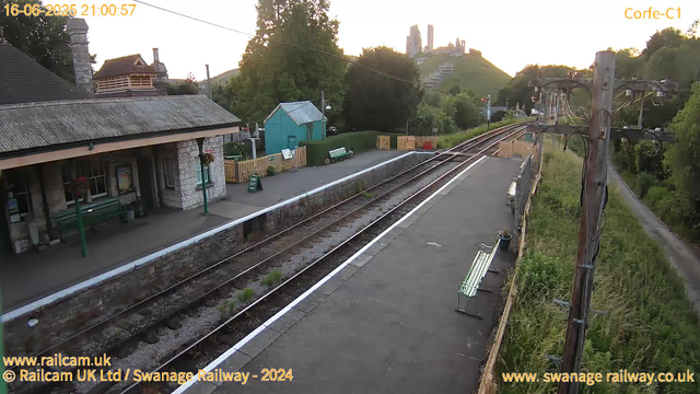 A small train station platform with a stone building on the left, featuring a chimney and window boxes with flowers. There are benches and a green sign that says "Way Out." Behind the station, there's a small turquoise building with a sloped roof, surrounded by a wooden picket fence. Green trees and hills are in the background, with a castle-like structure on top of a hill in the distance. To the right, railway tracks run alongside the platform, with electric wires and poles overhead. A narrow dirt path runs parallel to the tracks on the far right. The sky is clear with the sun setting, casting a warm glow over the scene. The timestamp indicates it is 21:00:57 on June 16, 2025.