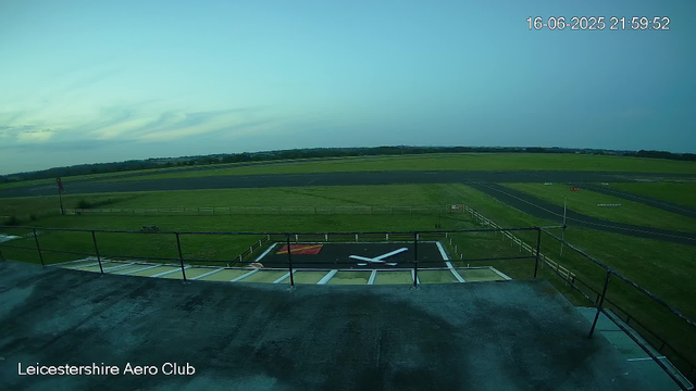 An outdoor view from a webcam at Leicesterhire Aero Club showing a grassy area with a runway in the background. In the foreground, there is a helipad with a large white "H" painted on a black surface, and a red square marked near the top left corner. The sky is partly cloudy with a calm atmosphere. The date and time displayed are 16-06-2025 at 21:59:52.